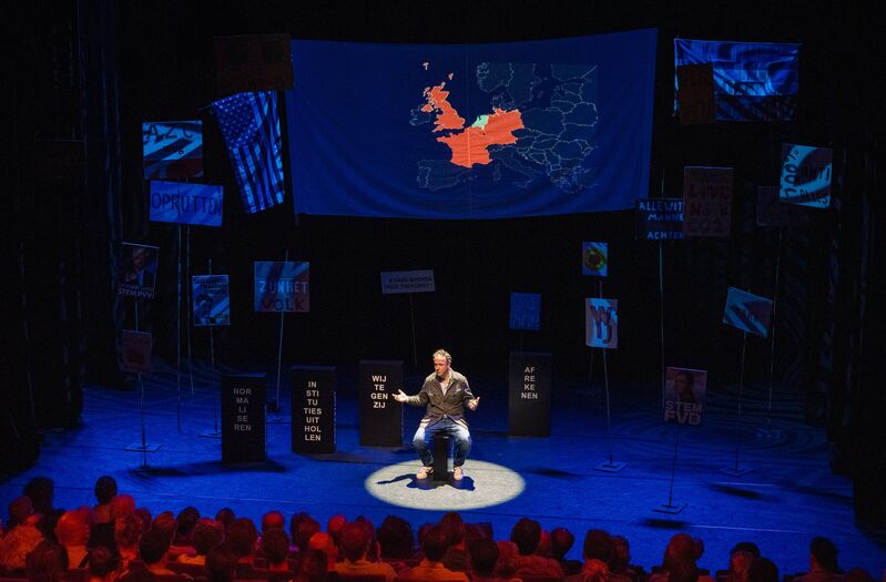 Een spreker op een podium in een theater met een kaart van Groot-Brittannië op de achtergrond en een publiek in het donker.