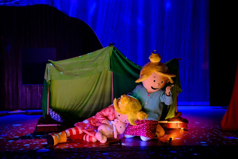 Een kleurige poppenvoorstelling met een meisje en een pop in een tent op een podium met zachte belichting.