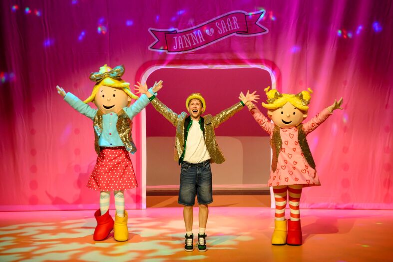 Drie vrolijke personages, waaronder een jongen en twee meisjes, staan op een podium en geven een show met hun armen omhoog.