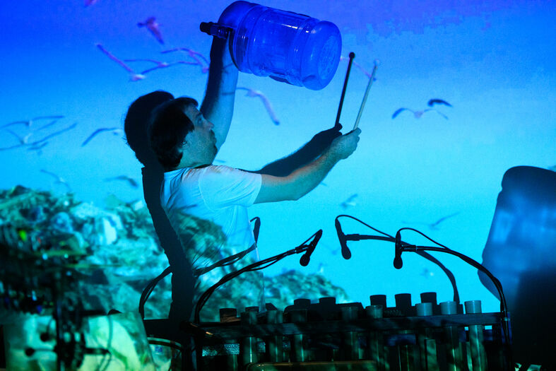 Een muzikant die een grote blauwe fles boven zijn hoofd houdt, met een kleurrijke achtergrond van visuele effecten.