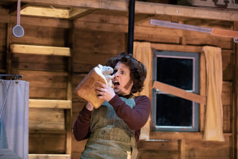 Acteur in kostuum houdt een groot stuk brood omhoog in een houten hut, met een raam op de achtergrond.