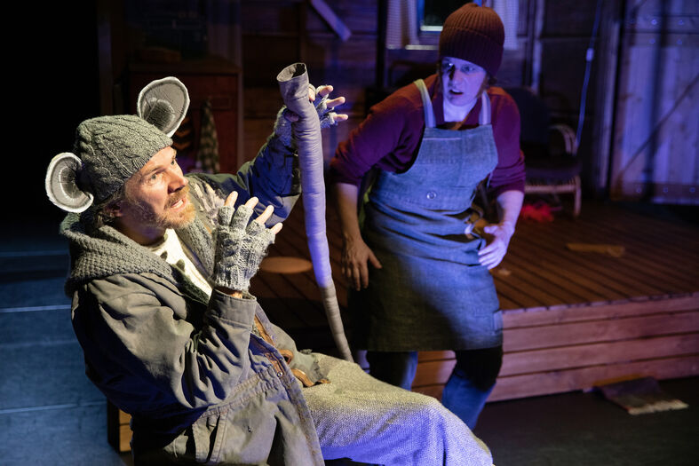 Een acteur verkleed als een koala zit op een stoel, terwijl een andere acteur toekijkt.