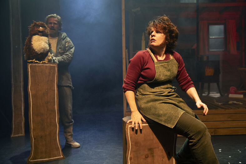 Twee acteurs in een theaterproductie; één met een pop, de ander zittend op een krat in een sfeervolle omgeving.