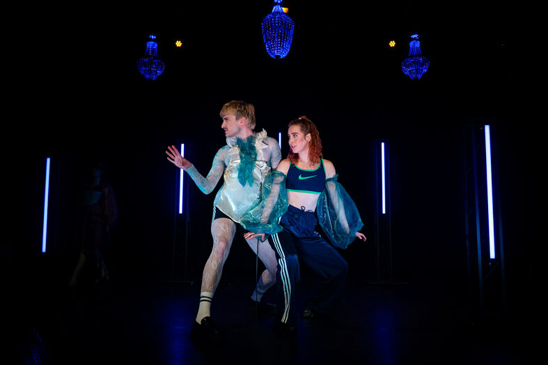 Twee performers in flamboyante kostuums dansen op een donker podium met sfeervolle verlichting.