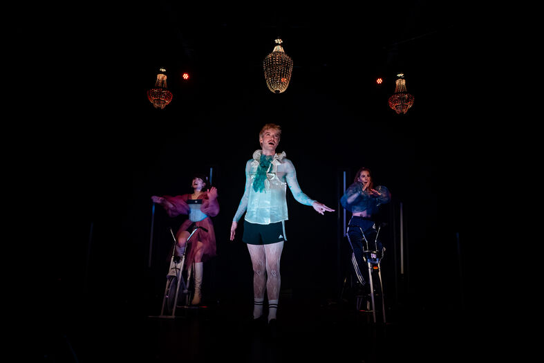 Drie performers in kleurige kostuums op een donkere podium, belicht door bovenliggende lampen.
