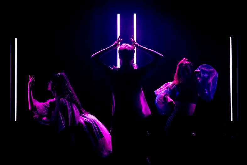 Drie mensen in silhouet, met neonlichten op de achtergrond, tijdens een dansperformance in een donkere omgeving.