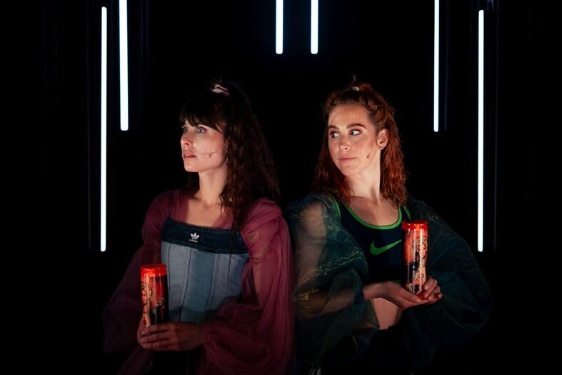 Twee vrouwen staan naast elkaar, elk met een kaars in de hand, tegen een donkere achtergrond met verticale lichtstralen.