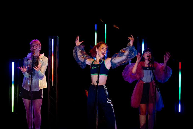 Drie vrouwelijke performers in kleurige verlichting, gekleed in moderne outfits, voeren een energieke act op.