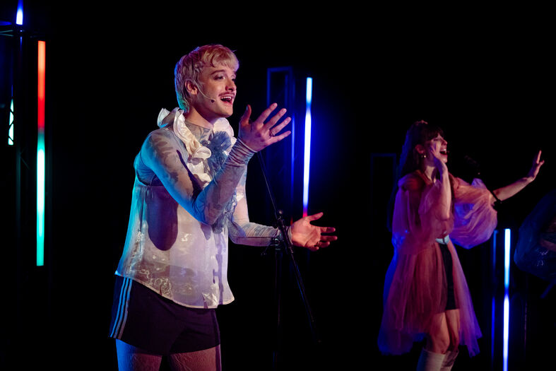 Twee performers op een podium met kleurrijke lichten; een zingt en de ander acteert.