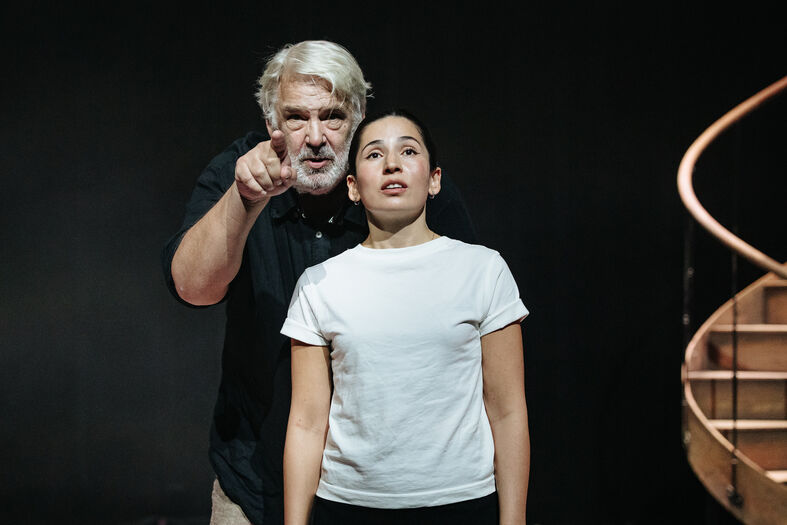 Een oudere man wijst boos naar een jongere vrouw, die geschokt en stil lijkt te staan in een donkere omgeving.