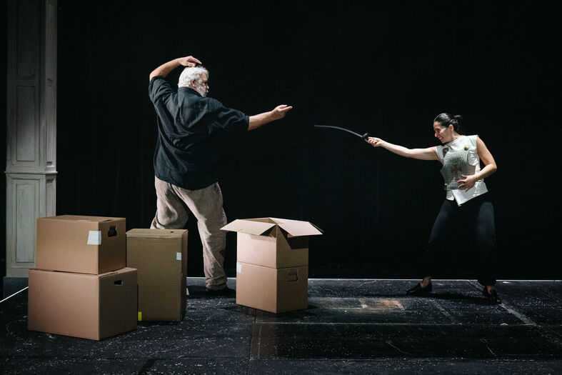Acteurs in een theatrale scène vechten met een zwaard en een stok, omgeven door kartonnen dozen.