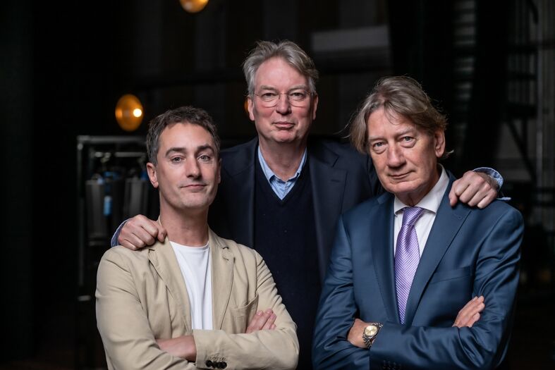 Drie mannen poseren samen voor een camera, met een neutrale achtergrond en lichte verlichting.