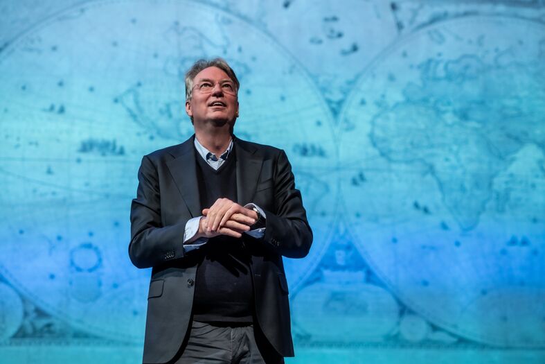 Een spreker staat op een podium met een achtergrond van wereldkaarten en presenteert zijn ideeën.