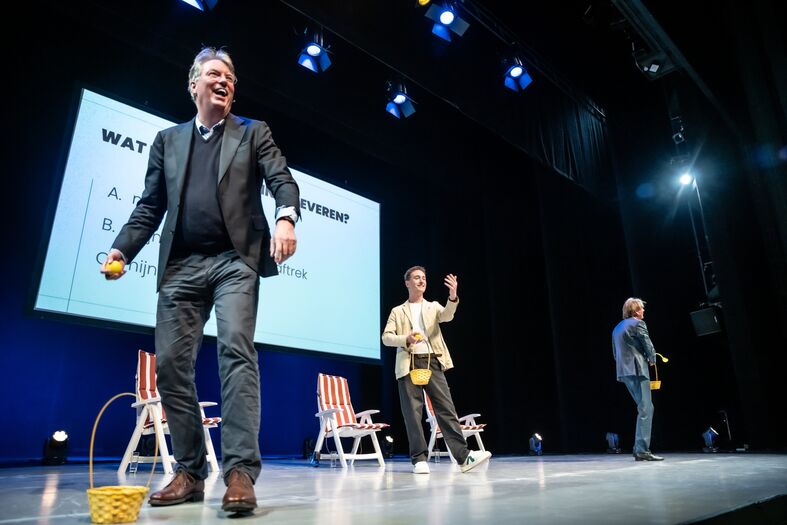 Drie mannen op een podium tijdens een presentatie, met een scherm op de achtergrond en verschillende attributen in hun handen.