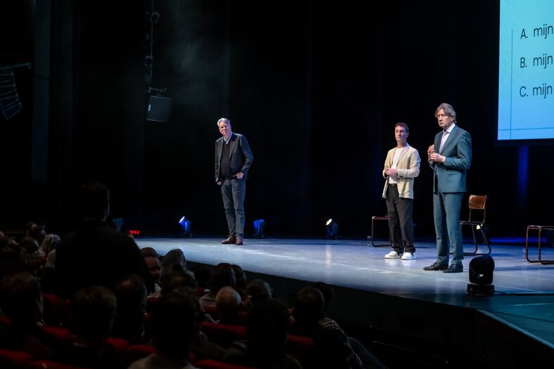 Twee sprekers staan op een podium voor een publiek, met stoelen en een projectiescherm op de achtergrond.