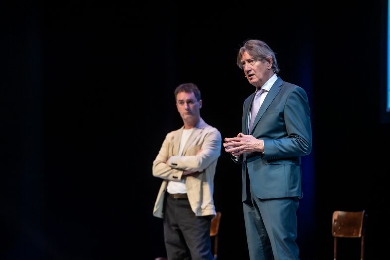 Twee mannen op een podium, één in een blazer en de ander in een lichte trui, terwijl ze met elkaar in gesprek zijn.
