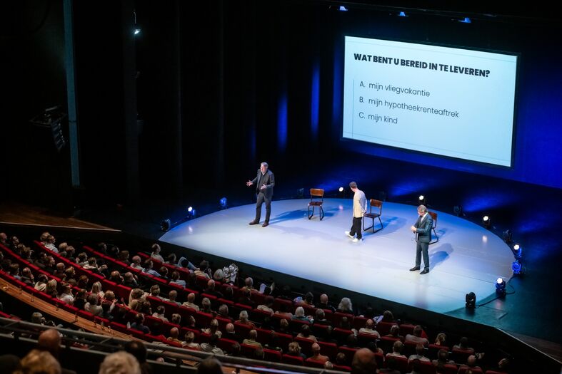 Een podium met sprekers tijdens een presentatie voor een publiek in een theater. Achtergrond met projectiescherm.