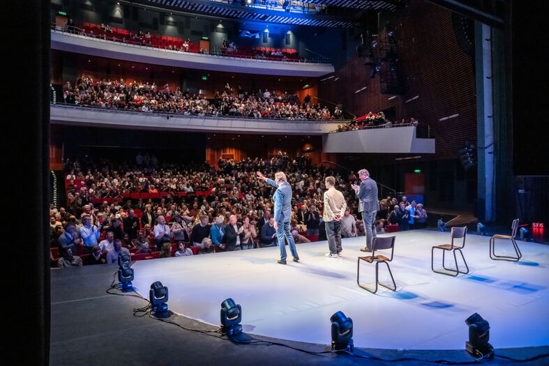 Drie performers staan op een podium met een volle theaterzaal in de achtergrond. Verlichting benadrukt de actie.