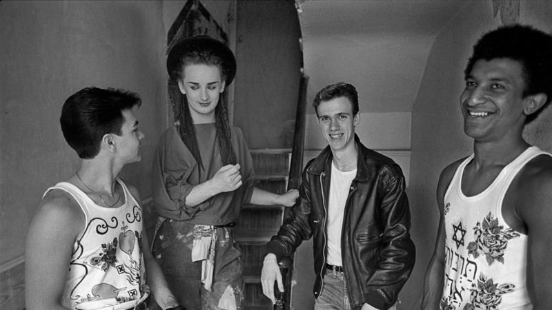 Groep van vier mensen, elk met verschillende stijlen, staan in een gang en lachen naar de camera. Monochrome afbeelding.