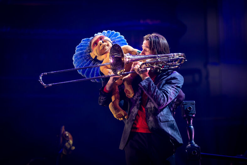 Performende artiest speelt op een trombone, terwijl hij een marionet in de vorm van een hond vasthoudt.