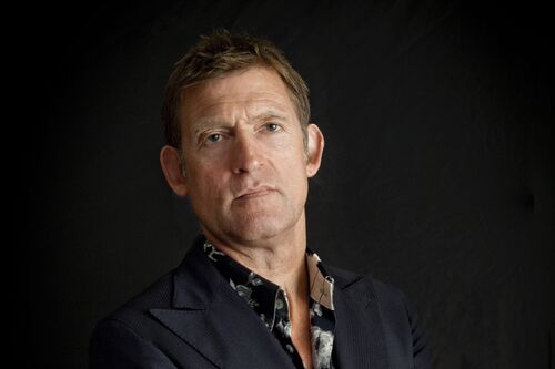 Portret foto van Huub van der Lubbe in een een pak met blouse met motief.