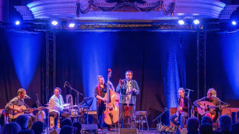 Zes muzikanten optreden op een podium met blauwe verlichting, bespelend verschillende instrumenten.