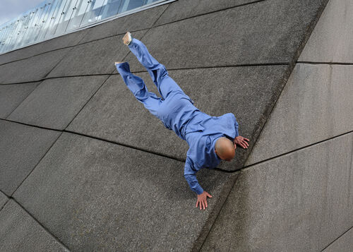 Een man in een blauwe outfit hangt ondersteboven aan een verticale muur, terwijl hij met zijn handen steun zoekt.