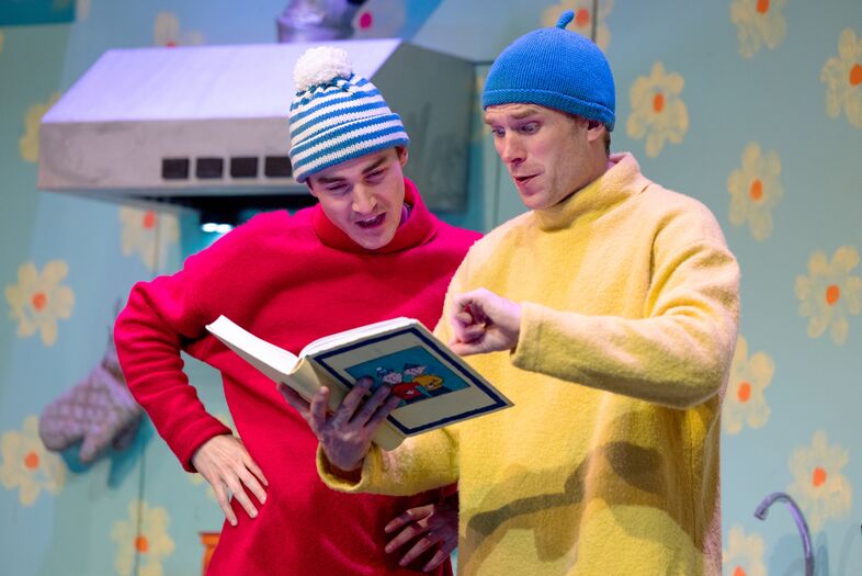 Twee acteurs in kleurrijke kostuums staan op een podium en lezen samen uit een groot boek.