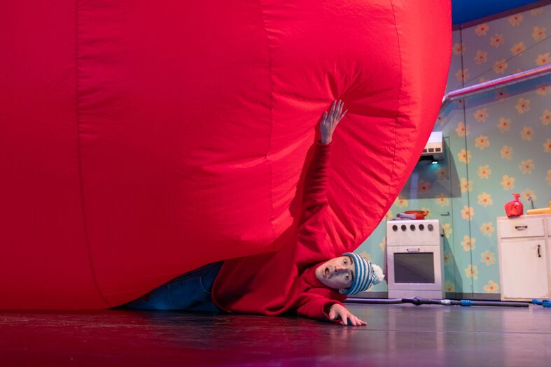 Een persoon zit gedeeltelijk onder een grote, rode ballon terwijl ze naar de camera kijkt in een kleurrijke ruimte.