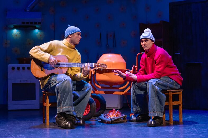 Twee acteurs in kleurrijke outfits zitten op stoelen op een podium, met een gitaar en een barbecue als decor.