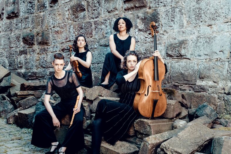 Vrouwenensemble poseert tussen stenen, met een cello en verschillende instrumenten, in een stijlvolle setting.