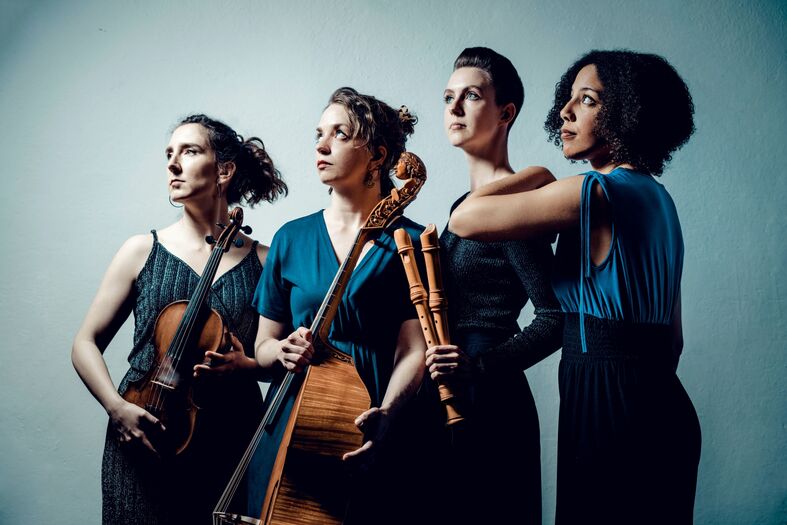 Vier vrouwelijke muzikanten poseren met verschillende muziekinstrumenten in een neutrale setting.