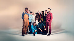 Grote groep personen in kleurrijke kleding poseert samen in een studio‑opstelling tegen lichte achtergrond.