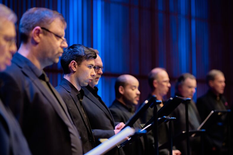Zanggroep bestaande uit tien mannen in zwart tenue, zittend op een podium met bladmuziek, onder blauwe verlichting.