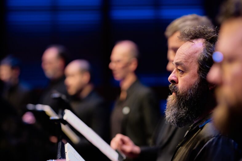 Een groep mannen met muziekpartituren, zittend op het podium tijdens een zanguitvoering.