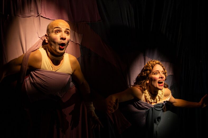 Twee acteurs in theatrale kostuums, emotioneel expressief, staan op een toneel met kleurrijke achtergronden.