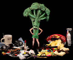 Grote broccoli‑figuur met menselijke armen en benen staat tussen verspreide etenswaren, snoep, kopje, chips en andere voorwerpen op een zwarte achtergrond.