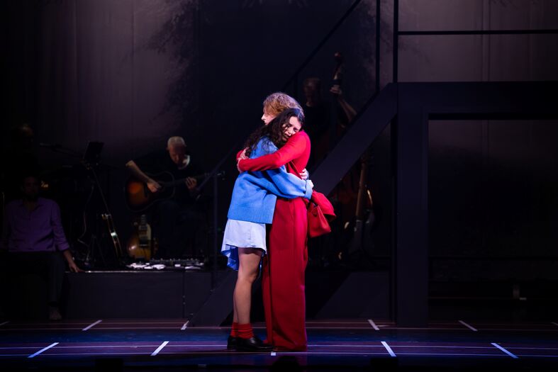Een emotioneel moment tussen twee acteurs die elkaar omarmen op het podium, met een muzikale achtergrond.