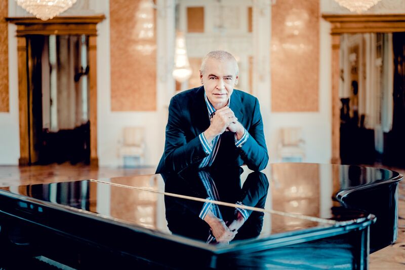 Een man in een zwart pak leunt op een piano in een elegante ruimte met luxe inrichting.
