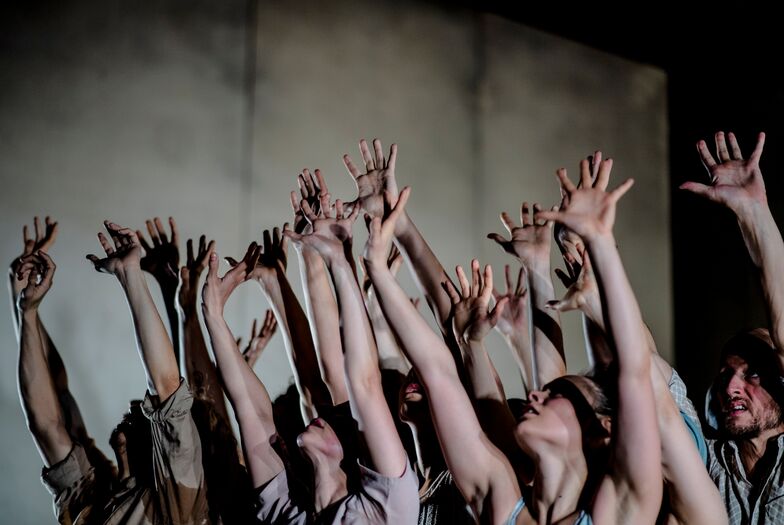 Groep mensen met opgestoken handen, in een theatrale setting met donkere achtergronden en dramatische belichting.