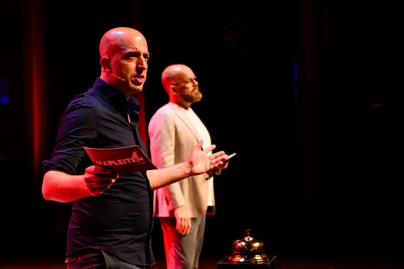 Twee mannen staan op een podium; de linker man spreekt met een microfoon, de rechter man houdt papier vast.