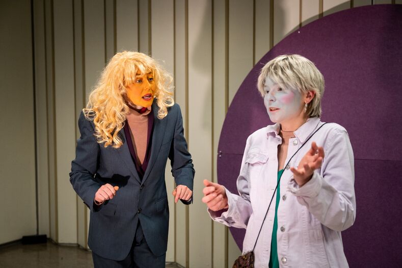 Twee acteurs op een podium in kostuums, één met een blond pruik en de ander met een neutrale make-up, in een dramatische scène.