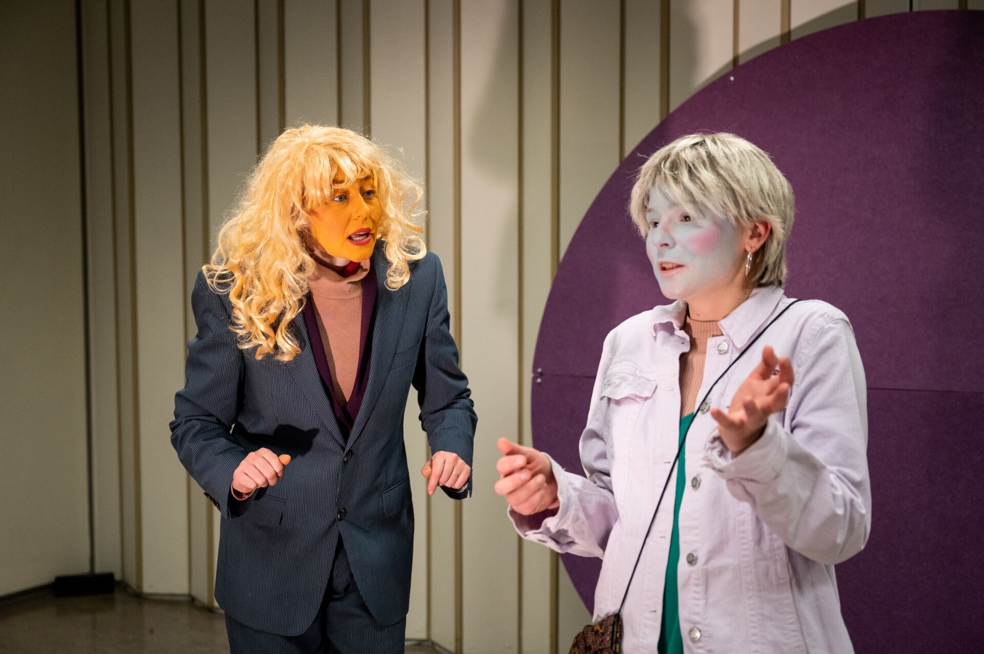 Twee acteurs op een podium in kostuums, één met een blond pruik en de ander met een neutrale make-up, in een dramatische scène.
