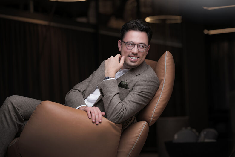 Een man in een stijlvolle outfit leunt ontspannen in een moderne leren stoel met een glimlach.