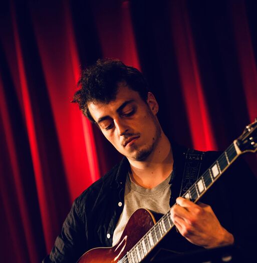 Man speelt gitaar op het podium met een rode achtergrond, geconcentreerd op zijn spel.