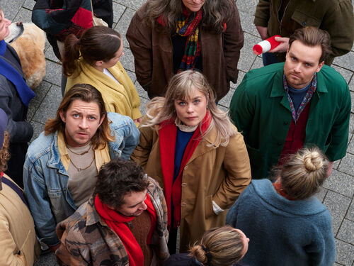 Groep mensen staat dicht bij elkaar op een plein en 3 personen kijken omhoog naar de camera