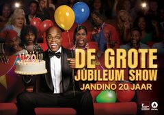 Groep personen zit op een podium met verjaardagstaart en gekleurde ballonnen tegen donkere achtergrond.