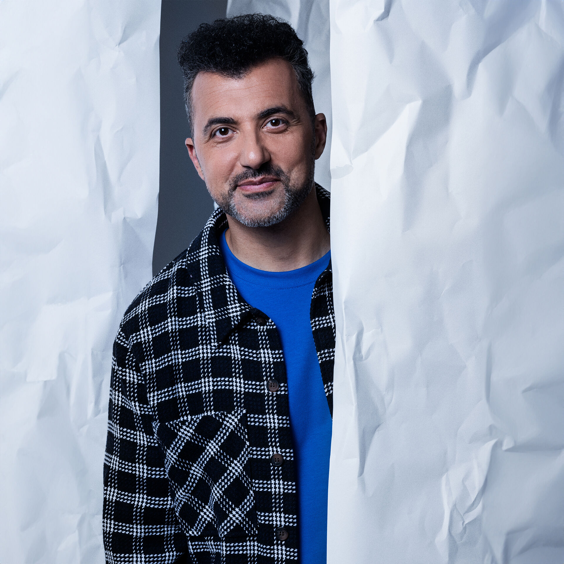 Man met een zwarte en witte geblokte overhemden en blauwe T-shirt, staat lachend tussen twee grote vouwen van wit papier.