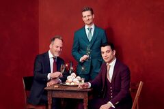 Drie personen in nette pakken zitten en staan rond een houten tafel met oliebollen en sterretjes tegen een rode achtergrond.
