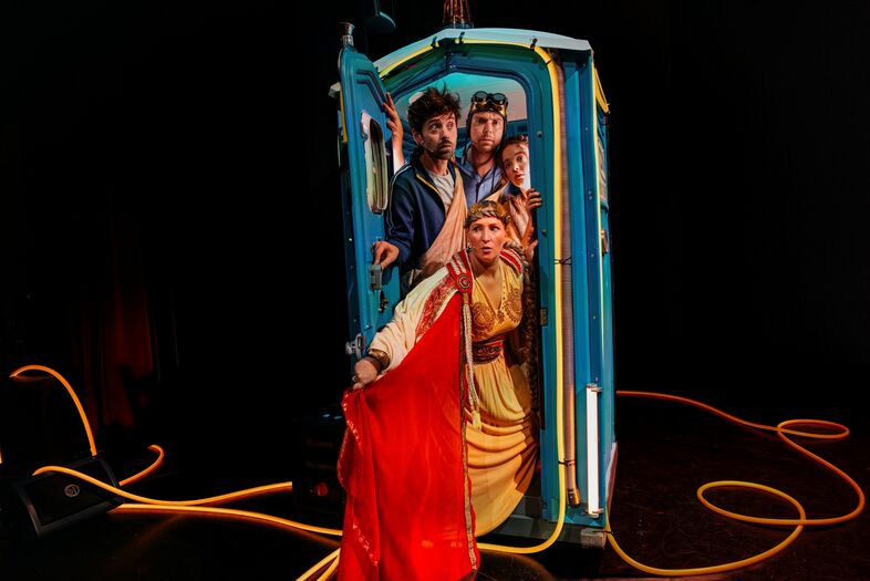 Drie mensen gekleed in kleurrijke kostuums staan in een blauw toilet met kabels op de vloer.